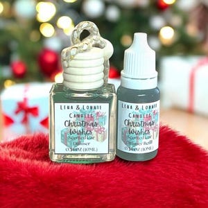May include: Two car air fresheners with the text "Christmas Wishes" are displayed on a red surface. One is a square glass bottle with a wooden top, and the other is a refill bottle. Both are labeled "Lena & Lonnie Candles".