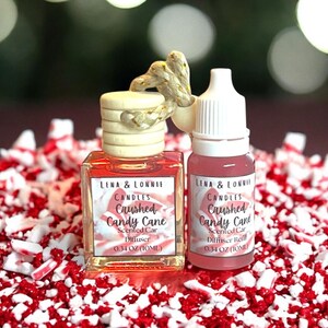 May include: Two car air fresheners with "LENA & LONNIE CANDLES Crushed Candy Cane" labels. One is a square bottle with a wooden top and rope detail, the other a refill bottle. Both contain 0.34 oz (10ml).