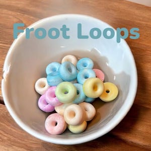May include: A white ceramic bowl containing a collection of small, ring-shaped cereal pieces in pastel shades of blue, pink, yellow, and white. The text Froot Loops is overlaid in light blue letters at the top of the image above the bowl.