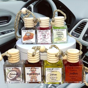 May include: Eight small glass bottles with wooden tops and string hangers. Each bottle contains a scented car diffuser with a different scent. The scents are Pumpkin Pie, Candy Cane, Cucumber Melon, Coconut, Gingerbread, Lemon, Leather, and a scent that is not visible in the image. The bottles are arranged in two rows of four. The bottles are on a white surface.