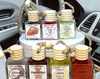 May include: Eight small glass bottles with wooden tops and string hangers. Each bottle contains a scented car diffuser with a different scent. The scents are Pumpkin Pie, Candy Cane, Cucumber Melon, Coconut, Gingerbread, Lemon, Leather, and a scent that is not visible in the image. The bottles are arranged in two rows of four. The bottles are on a white surface.