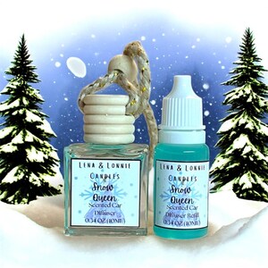 May include: A car air freshener set with a winter theme. The set includes a square diffuser bottle with a wooden top and a refill bottle. Both bottles have labels that read "Snow Queen" and "Scented Car Diffuser". The background features snowy pine trees.