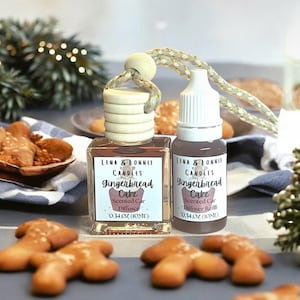 May include: A car air freshener and refill set with gingerbread cake scent. The set includes a square glass diffuser bottle with a wooden top and a small dropper bottle. Both bottles have labels with the text "Lena & Lonnie Candles Gingerbread Cake". Gingerbread cookies are scattered around.