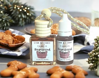 May include: A car air freshener and refill set with gingerbread cake scent. The set includes a square glass diffuser bottle with a wooden top and a small dropper bottle. Both bottles have labels with the text "Lena & Lonnie Candles Gingerbread Cake". Gingerbread cookies are scattered around.