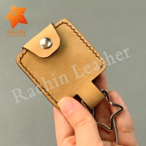 May include: A tan leather keychain holder with a star-shaped keyring. The holder is square with a flap fastened by a silver stud. The text "Rachin Leather" is visible on the holder.
