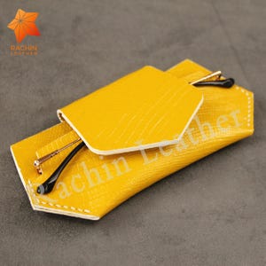 Może przedstawiać: Żółte skórzane etui na okulary z białymi szwami. Etui jest złożone i zamknięte, a wewnątrz znajdują się okulary. Na etui widoczny jest napis "Rachin Leather".
