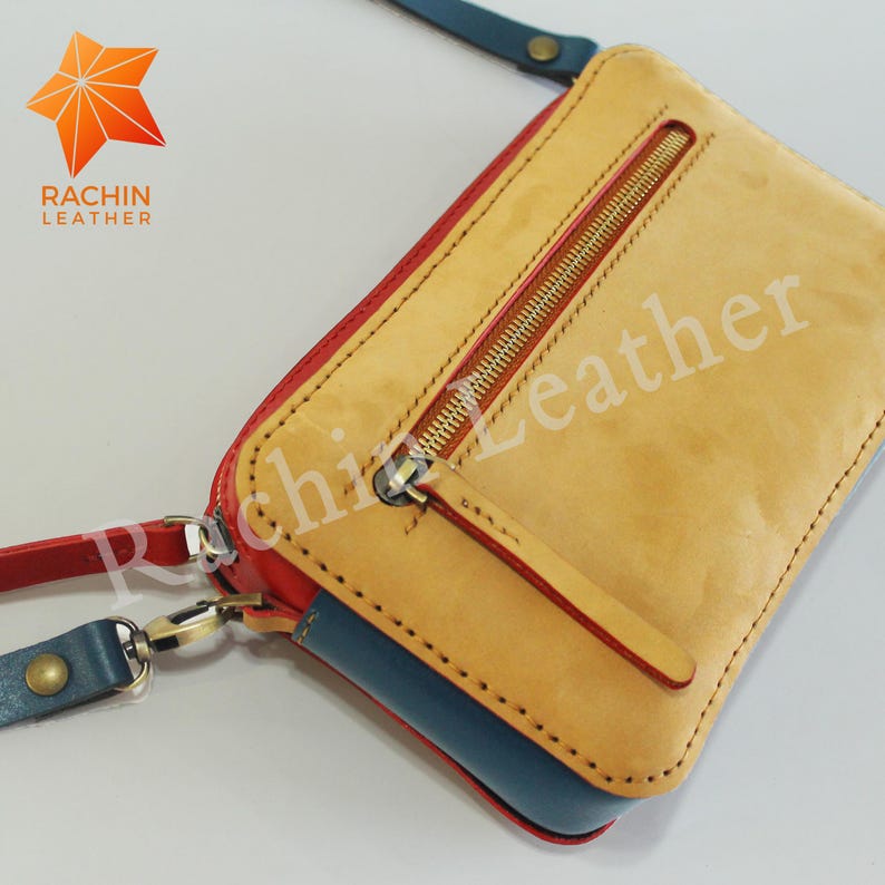 May include: A small, tan leather purse with a red trim and a zipper closure. The purse has a blue leather lining and a gold zipper pull. The purse is made by Rachin Leather.