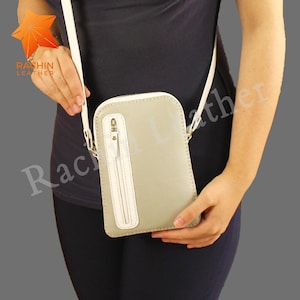 May include: A light gray leather crossbody bag with a white zipper and strap. The bag is rectangular with rounded corners and is being held by a person. The brand name "RASHIN LEATHER" is visible on the upper left.