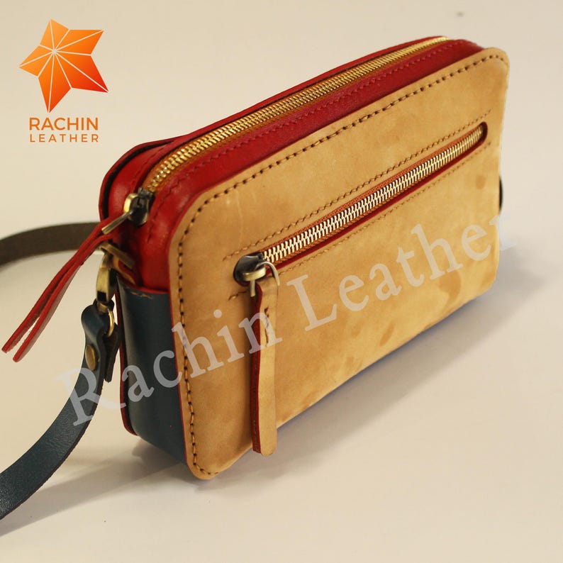 May include: A small, tan leather crossbody bag with a red and blue trim. The bag has two gold zippers and a gold zipper pull. The bag is labeled "Rachin Leather".