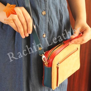 May include: A small, tan leather crossbody bag with a red zipper and a blue leather flap. The bag has a gold zipper pull and a gold clasp. The bag is being held by a person's hand.