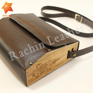 Peut inclure: Un sac à bandoulière en cuir marron avec un rabat en bois orné d'un motif floral gravé. Le sac est doté d'une bandoulière en cuir marron et d'une boucle en métal.
