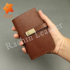 Könnte beinhalten: Ein braunes Lederportemonnaie mit strukturierter Oberfläche und einem messingfarbenen Verschluss. Das rechteckige Portemonnaie hat einen Sicherungsriemen. Der Markenname "Rachin Leather" ist auf dem Portemonnaie aufgedruckt.