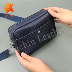 May include: A navy blue leather crossbody bag with a zippered top and a front pocket with a snap closure. The bag has a matching blue adjustable strap and silver-tone hardware. The brand name "Rachin Leather" is visible on the bag.