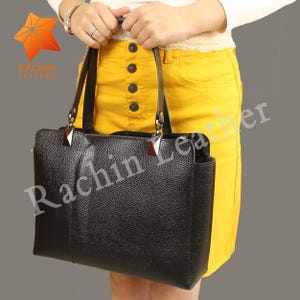 Puede incluir: Bolso tote de cuero negro con acabado texturizado, forma rectangular y asas superiores dobles. El bolso tiene herrajes plateados y un diseño estructurado. El nombre de la marca "Rachin Leather" es visible.