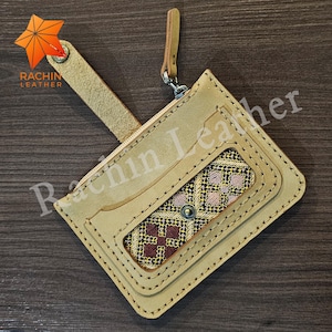 May include: A light tan leather wallet with a patterned fabric insert. The wallet has a zipper closure, a wrist strap, and a key ring. The text "Rachin Leather" is visible on the wallet.