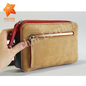 May include: A tan leather clutch bag with a red zipper and a gold zipper pull. The bag has a small pocket on the front with a red zipper. The bag is made by Rachin Leather.
