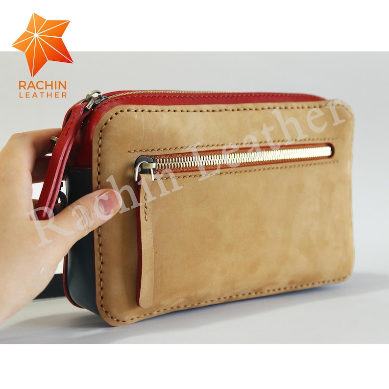 May include: A tan leather clutch bag with a red zipper and a gold zipper pull. The bag has a small pocket on the front with a red zipper. The bag is made by Rachin Leather.