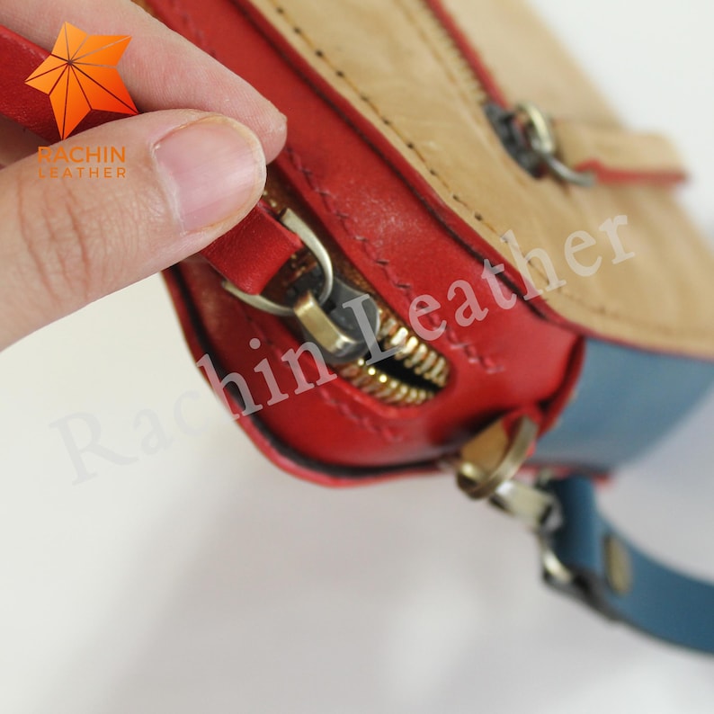 May include: Close-up of a red, blue, and tan leather bag with a gold zipper. The bag has a small strap and a zipper pull with a metal ring.