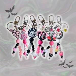 Könnte beinhalten: Eine Sammlung von Schlüsselanhängern zum Thema Halloween mit verschiedenen Anhängern, darunter Totenköpfe, Geister und Herzen. Jeder Schlüsselanhänger hat eine Mischung aus rosa, schwarzen und weißen Perlen mit silberfarbenen Beschlägen. Einige Anhänger haben den Text "Stay Spooky" und "Call Me".