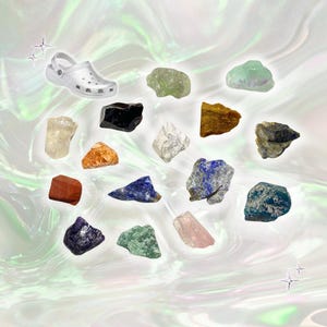 May include: A collection of various colorful raw gemstones and a silver clog shoe are arranged on a pearlescent background. The gemstones include shades of green, blue, orange, red, and purple. The shoe is a classic style.