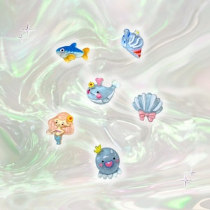 May include: A collection of colorful, cartoon-style ocean-themed charms. The charms include a blue and yellow fish, a blue and white seashell, a blue whale with a crown, a mermaid, a blue octopus with a crown, and a seashell with a pink bow.