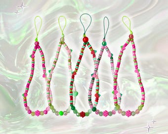 May include: Five beaded phone charms in shades of pink, green, and red. Each charm features a loop at the top and is adorned with various beads, including small strawberries and flower-shaped beads. The charms are displayed against a soft, iridescent background.