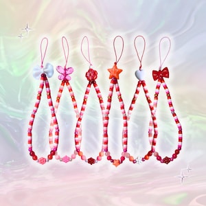 Peut inclure: Collection de cinq breloques de téléphone perlées en rouge, rose et blanc. Chaque breloque présente un élément décoratif différent : un nœud, un papillon, une fleur, une étoile, un cœur et un autre nœud. Les breloques sont enfilées sur un cordon rose.