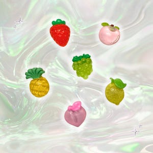 May include: A collection of colorful fruit-shaped charms, including a red strawberry, a green bunch of grapes, a yellow pineapple, a pink peach, a yellow lemon, and a pinkish-red fruit. Each charm has detailed features and a glossy finish.