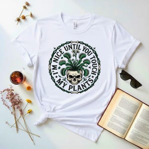 May include: A white short-sleeve t-shirt featuring a circular graphic of a human skull acting as a planter for green Venus flytrap plants. The design is encircled by thorny vines and the text I'M NICE UNTIL YOU TOUCH MY PLANTS in black.