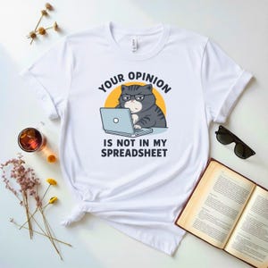 Pode incluir: Uma t-shirt branca de manga curta com o gráfico de um gato cinzento tigrado a usar óculos e a digitar num portátil. O texto acima e abaixo do gato diz "YOUR OPINION IS NOT IN MY SPREADSHEET" sobre um fundo circular amarelo.