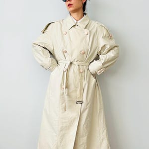 Op de afbeelding: Een beige trenchcoat met een dubbele rij knopen, grote knopen en een bijpassende riem. De jas heeft een klassiek silhouet met een kraag en lange mouwen. De jas is ongeveer 140 cm lang.