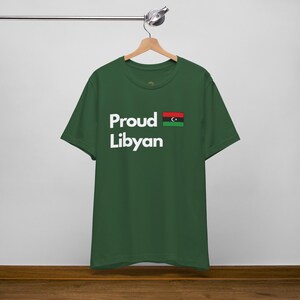Può includere: Una maglietta verde scuro con il testo "Proud Libyan" in lettere bianche e una piccola bandiera libica stampata sul lato destro del petto.