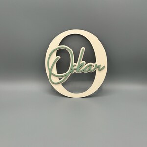 May include: A wooden cutout of the letter "O" with the name "Oskar" written in a cursive font inside the letter. The letter is painted a light beige color and the name is painted a light green color.