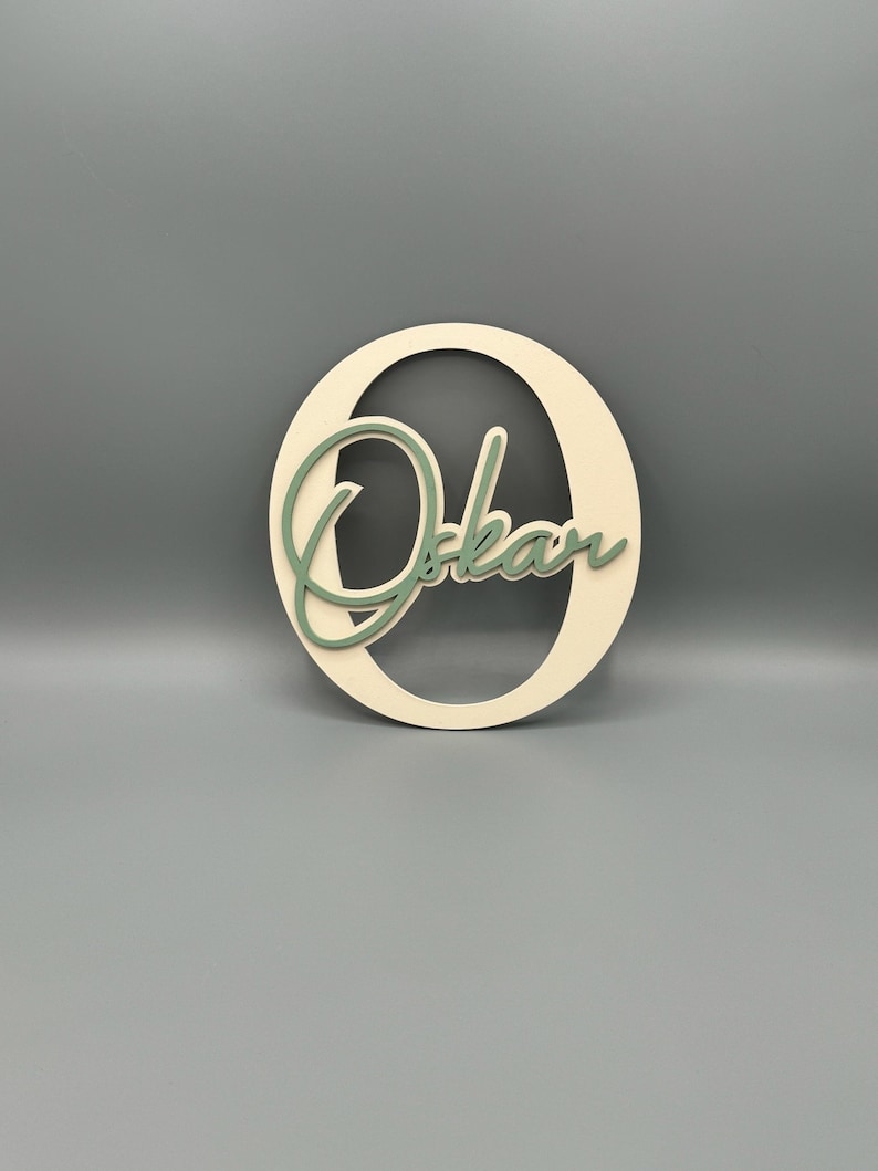 May include: A wooden cutout of the letter "O" with the name "Oskar" written in a cursive font inside the letter. The letter is painted a light beige color and the name is painted a light green color.