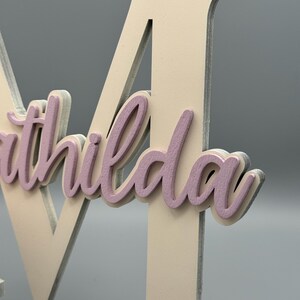 May include: A white wooden letter M with the name "Mathilda" written in pink on it.