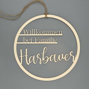 May include: A wooden circle with the text "Willkommen bei Familie Harbauer" in a cursive font. The circle is hanging from a twine rope.