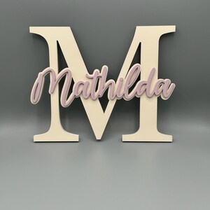 May include: A white wooden letter M with the name "Mathilda" cut out in a light pink color.