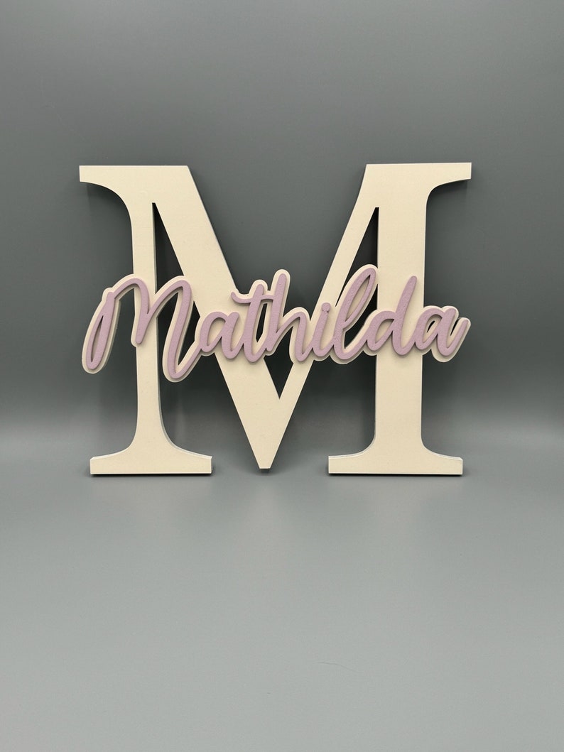 May include: A white wooden letter M with the name "Mathilda" cut out in a light pink color.