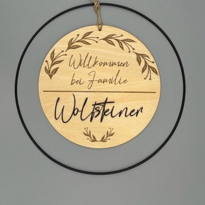 May include: A round wooden sign with the text "Willkommen bei Familie Woltsteiner" and a floral design. The sign is hanging from a black metal ring.