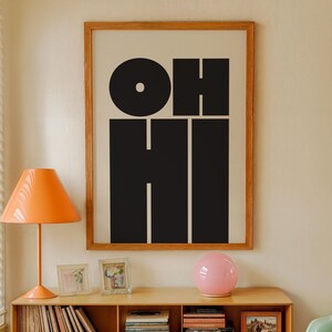 Könnte beinhalten: Ein Wohnzimmer im Mid-Century-Modern-Stil mit einem Holzschrank, Büchern und einem gerahmten Druck mit der Aufschrift "OH HI". Es gibt eine orangefarbene Lampe, eine rosa Kugellampe und einen hellgrünen Stuhl.