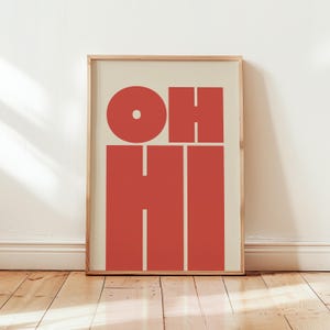 May include: A framed art print with the words "OH HI" in a bold, red font against a cream background. The letters are large and blocky, with a simple, modern design. The frame is a light wood color.