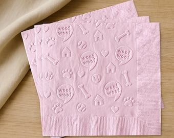 May include: Pink paper napkins with an embossed pattern of dog-themed icons, including paw prints, bones, houses, and speech bubbles with the text "Woof Woof". The napkins are stacked on a wooden surface.