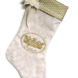 May include: A Christmas stocking with a white and gold floral pattern. The cuff has a gold chevron design. An embroidered oval features a gold thistle design and the word "Scotland". A white flower and ribbon are attached.