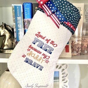 May include: A patriotic Christmas stocking with a white background and small gray polka dots. The top is navy blue with white stars. The text "Land of the Free because my daddy is BRAVE" is embroidered on the front.