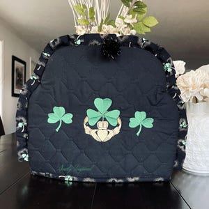May include: A quilted black tea cozy with a diamond pattern, featuring embroidered green shamrocks and a gold Claddagh design. The edges are trimmed with a patterned fabric. The text "Sewly Inspired" is visible on the bottom.