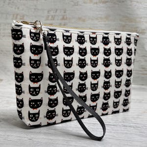 May include: A white zippered pouch with a repeating pattern of black cat faces. The cats have various expressions and some wear bow ties. The pouch has a gold zipper and a black wrist strap.