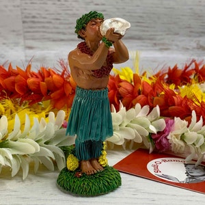 May include: A figurine of a man blowing a conch shell, wearing a green skirt and a lei. The figurine is on a green base with yellow flowers. Colorful leis in orange, white, and yellow are in the background.