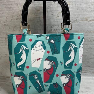 K&ouml;nnte beinhalten: Eine t&uuml;rkisblaue Tragetasche mit einem schwarzen Bambusgriff und goldfarbener Hardware. Die Tasche hat ein sich wiederholendes Muster von Charakteren in S&auml;rgen mit roten Akzenten. Die Tasche steht auf einem schwarzen St&auml;nder.