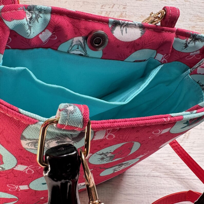 Puede incluir: Un bolso tote rojo con un interior turquesa y un exterior estampado con ilustraciones turquesas y blancas. El bolso tiene una correa de cadena dorada y un cierre negro.