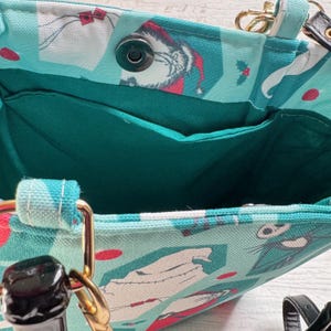 K&ouml;nnte beinhalten: Eine tealfarbene Tragetasche mit einem festlichen Grinch-Motiv. Die Tasche hat einen Magnetverschluss und eine dunkelgr&uuml;ne Innentasche. Goldfarbene Beschl&auml;ge akzentuieren die Griffe und Befestigungen der Tasche.